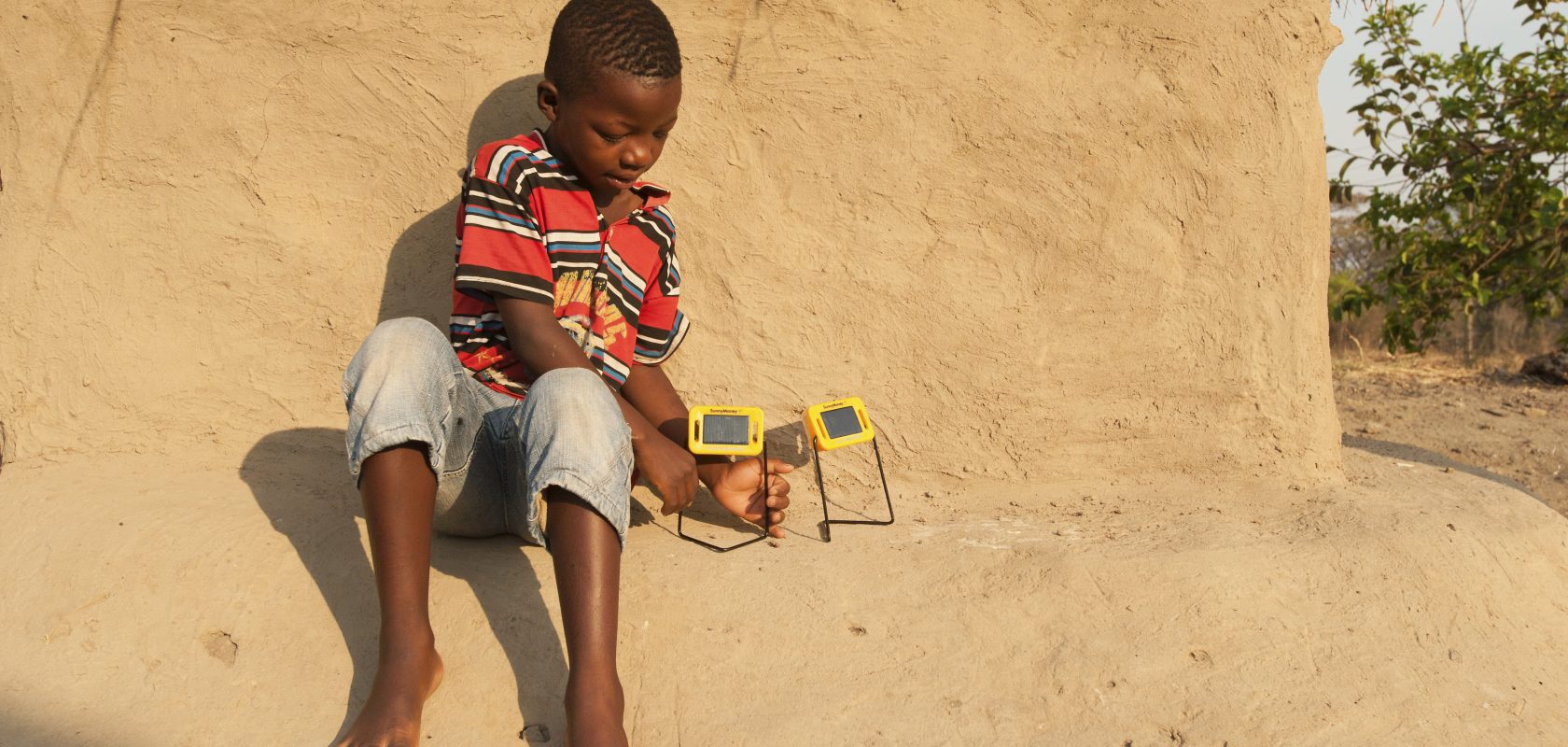 Young boy sits on the side of a mud hut while charging his solar lights.