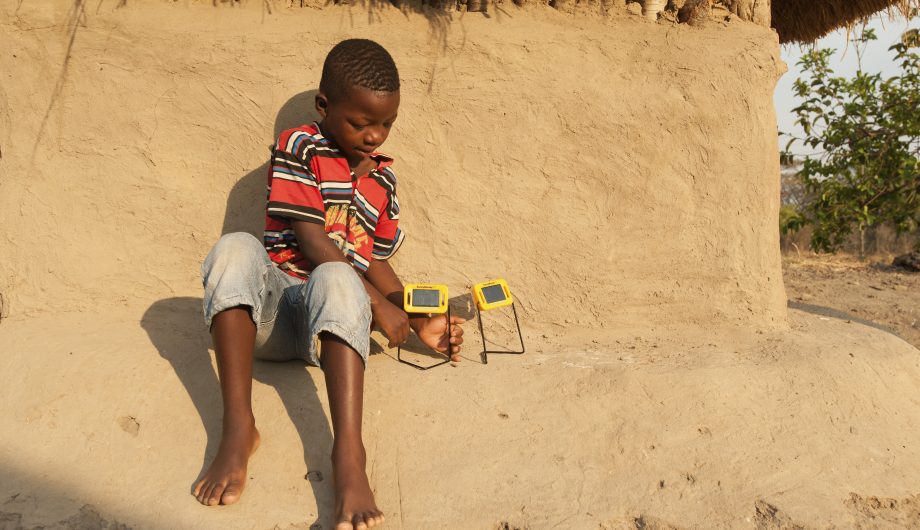Young boy sits on the side of a mud hut while charging his solar lights.