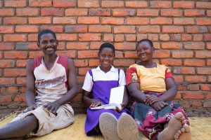 A family sits on a straw mat in front of a brick wall.