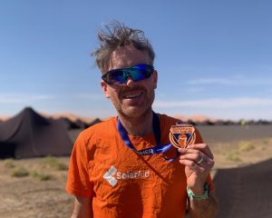 Man holds medal from Marathon des Sables in the desert.