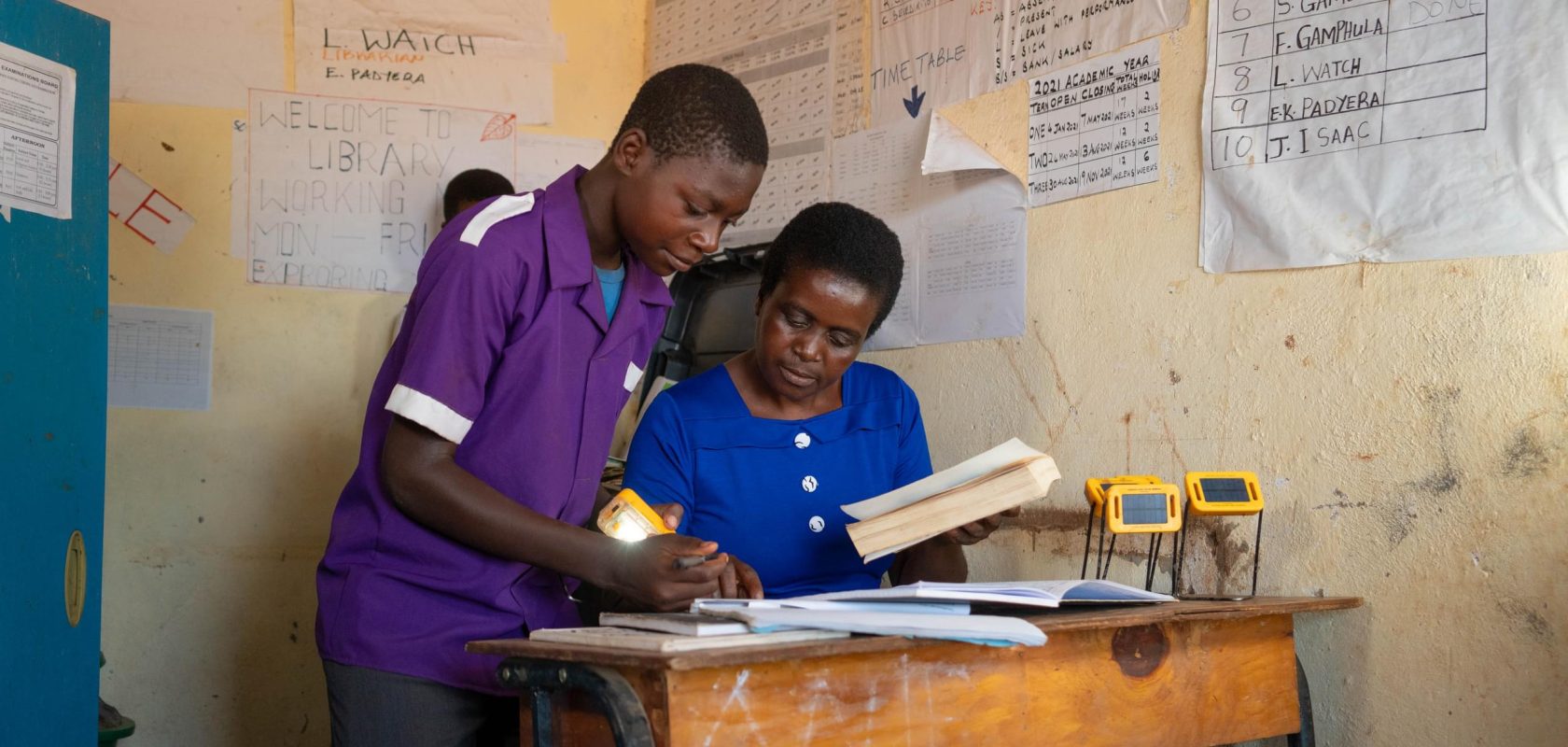 Dalitso stands with his school teacher Beatrice as they look at his work. There are solar lights on the desk.