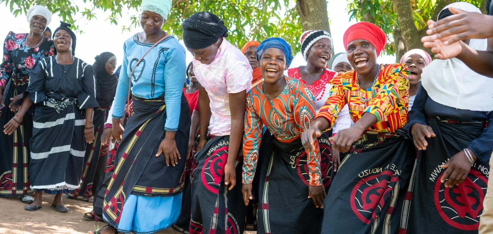 A group of women from the Chimwemwe Mayi Walas are dancing and celebrating together under a tree.