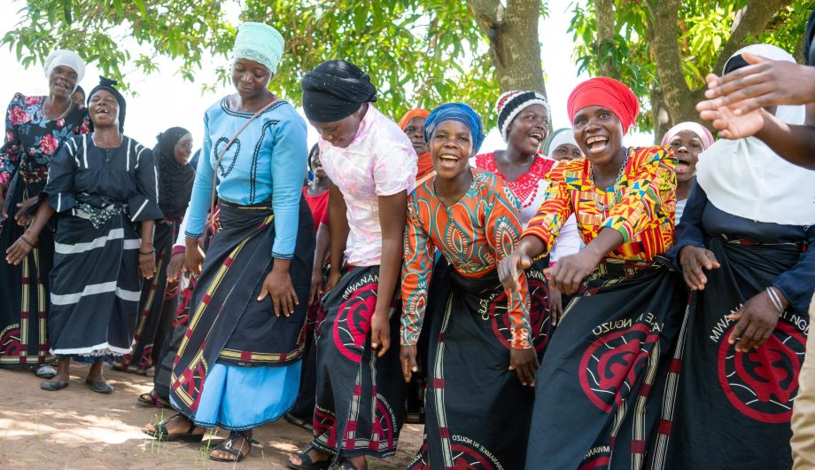 A group of women from the Chimwemwe Mayi Walas are dancing and celebrating together under a tree.